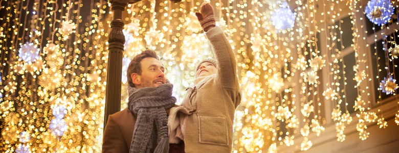 A father holds his young daughter in his arms as she reaches out towards hanging Christmas lights