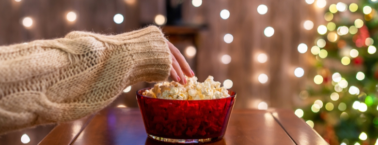 A woman eating popcorn and watching TV in a room decorated for Christmas.