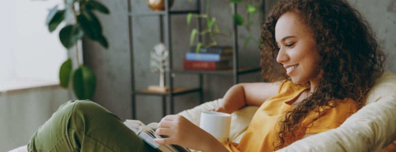 A woman sitting in a chair while drinking a cup of tea and reading a book.