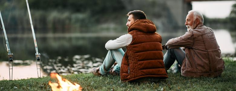 Two men sitting together and fishing by a river.